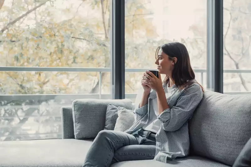 A smiling woman holding a cup of coffee or tea, sitting on a couch and looking out the window.