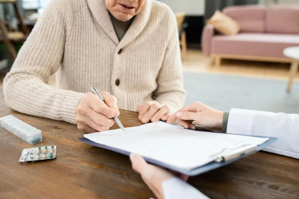 Sick senior man sitting at a table with a doctor, filling out information on a clipboard with prescription nearby